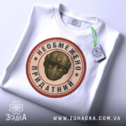 Футболка необмежено придатний, білого кольору, складена на поверхні, фон світло-сірий, принт у центрі з зображенням шолома та написом, прикріплена етикетка з зеленим шнурком.