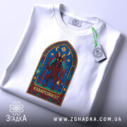 Футболка Ніч Крампуса, біла, зображена зверху, на фоні світло-сірого градієнту, принт яскравих кольорів з Крампусом в арковій рамці, з етикеткою на комірці.