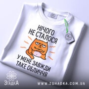 Футболка нічого не сталось. Біла футболка, складена на поверхні, з видимим етикеткою. Принт з оранжевим котом, текст чорного кольору: 'НІЧОГО НЕ СТАЛОСЯ У МЕНЕ ЗАВЖДИ ТАКЕ ОБЛИЧЧЯ'. Фон світло-сірий.