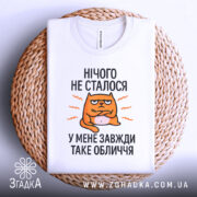Футболка нічого не сталось. Біла футболка, складена на плетеній основі. Принт з оранжевим котом, текст чорного кольору: 'НІЧОГО НЕ СТАЛОСЯ У МЕНЕ ЗАВЖДИ ТАКЕ ОБЛИЧЧЯ'. Фон світло-сірий.