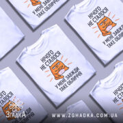 Футболка нічого не сталось. Біла футболка з коротким рукавом, зображена на манекені. Принт з оранжевим котом, який сидить з закритими руками, текст чорного кольору: 'НІЧОГО НЕ СТАЛОСЯ У МЕНЕ ЗАВЖДИ ТАКЕ ОБЛИЧЧЯ'. Фон світло-сірий.