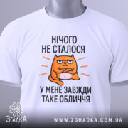 Футболка нічого не сталось. Біла футболка, складена на поверхні, з видимим етикеткою. Принт з оранжевим котом, текст чорного кольору: 'НІЧОГО НЕ СТАЛОСЯ У МЕНЕ ЗАВЖДИ ТАКЕ ОБЛИЧЧЯ'. Фон світло-сірий.