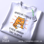 Футболка нічого не сталося. Біла футболка, складена на столі, з принтом жовто-оранжевого кота, що має незадоволений вираз обличчя, текст 'У МЕНЕ ЗАВЖДИ ТАКЕ ОБЛИЧЧЯ' під ним. Фон світло-сірий, на футболці є етикетка.