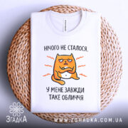 Футболка нічого не сталося. Біла футболка, складена на плетеному кошику, з принтом жовто-оранжевого кота, що виглядає незадоволеним, текст 'У МЕНЕ ЗАВЖДИ ТАКЕ ОБЛИЧЧЯ' під ним. Фон світло-сірий, футболка має округлий виріз.
