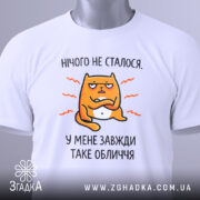Футболка нічого не сталося. Біла футболка, складена на столі, з принтом жовто-оранжевого кота, що має незадоволений вираз обличчя, текст 'У МЕНЕ ЗАВЖДИ ТАКЕ ОБЛИЧЧЯ' під ним. Фон світло-сірий, на футболці є етикетка.