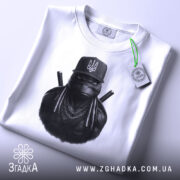 Футболка ніндзя український дух, біла, складена на столі, фон світло-сірий, принт з ніндзя в чорному капелюсі з гербом України, прикріплена етикетка з логотипом.