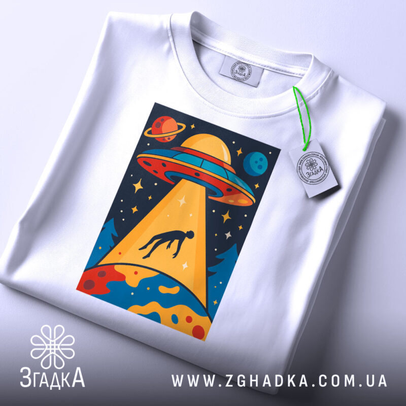 Футболка НЛО викрадення людини, біла, зображена зверху, фон світлий, принт з НЛО, що світить на людину, з яскравими кольорами та деталями, прикріплена етикетка з зеленим шнурком.