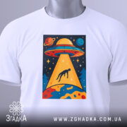 Футболка НЛО викрадення людини, біла, зображена зверху, фон світлий, принт з НЛО, що світить на людину, з яскравими кольорами та деталями, прикріплена етикетка з зеленим шнурком.