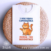 Футболка нормальна самооцінка. Біла футболка, складена на плетеній основі, з принтом, де зображений кіт оранжевого кольору. Текст на футболці синього та оранжевого кольорів. Фон світло-сірий, видно плетений елемент під футболкою.