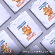 Футболка нормальна самооцінка. Біла футболка з принтом, на якому зображений усміхнений кіт оранжевого кольору. Принт розташований на грудях, текст синього та оранжевого кольорів. Фон світло-сірий, футболка представлена на манекені.