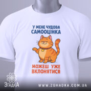 Футболка нормальна самооцінка. Біла футболка, складена на поверхні, з принтом, де зображений кіт оранжевого кольору. Текст на футболці синього та оранжевого кольорів. На футболці є етикетка з зеленим шнурком. Фон світло-сірий.