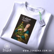 Футболка новий рік. Біла футболка, складена на поверхні, з принтом на чорному фоні, зображення жінки з ялинкою. На футболці є етикетка з зеленим шнурком.