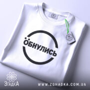 Футболка обнулись стильний мінімалізм, біла, складена на поверхні, фон світло-сірий, принт чорний з написом 'Обнулись' у колі, на комірці є етикетка.