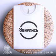 Футболка обнулись стильний мінімалізм, біла, складена на плетеній кошику, фон світло-сірий, принт чорний з написом 'Обнулись' у колі.