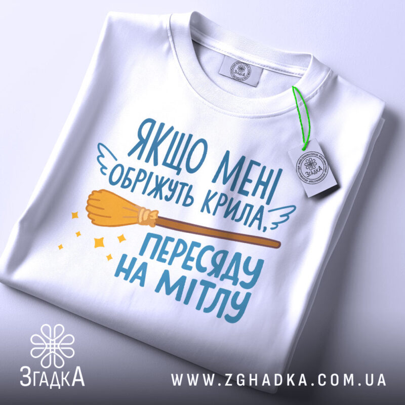 Футболка обріжуть крила, біла, складена на поверхні, зображена з верхнього ракурсу, фон світло-сірий, принт з текстом та малюнком мітли, на футболці є етикетка з логотипом.