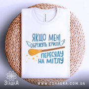 Футболка обріжуть крила, біла, складена на плетеній кошику, зображена з верхнього ракурсу, фон світло-сірий, принт з текстом та малюнком мітли, виглядає акуратно.