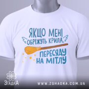 Футболка обріжуть крила, біла, складена на поверхні, зображена з верхнього ракурсу, фон світло-сірий, принт з текстом та малюнком мітли, на футболці є етикетка з логотипом.