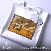 Футболка операція павутина, складена футболка білого кольору, фон світлий, принт з зображенням вантажівки на фоні пожежі та дронів, з етикеткою на шнурку.