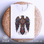Футболка орел бойовий дух, біла, зображена зверху, складена на плетеній кошику, фон світло-сірий, принт з орлом в червоних, чорних і золотих кольорах, деталі геометричні та символічні.