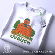 Футболка Овруч білого кольору, складена на поверхні, зображена з верхнього ракурсу, фон світло-сірий, принт з зображенням церкви з червоної цегли та зеленими деревами, напис OVRUCH під малюнком, на футболці є етикетка.