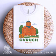 Футболка Овруч білого кольору, складена на плетеній основі, зображена з верхнього ракурсу, фон світло-сірий, принт з зображенням церкви з червоної цегли та зеленими деревами, напис OVRUCH під малюнком.