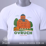 Футболка Овруч білого кольору, складена на поверхні, зображена з верхнього ракурсу, фон світло-сірий, принт з зображенням церкви з червоної цегли та зеленими деревами, напис OVRUCH під малюнком, на футболці є етикетка.