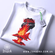 Футболка Палаючий літак, біла, зображена зверху, на фоні світлого градієнту. Принт з червоним літаком, що летить через полум'я, з текстом на чорному фоні, з зеленим ярликом.