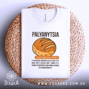 Футболка паляниця, білого кольору, зображена зверху на плетеній основі, фон нейтральний, принт з малюнком хліба та текстом українською мовою.