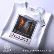 Футболка памʼять 24 лютого, біла, складена на поверхні, фон світлий, принт з палаючими будинками, на футболці є етикетка.