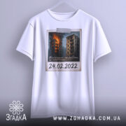 Футболка памʼять 24 лютого, біла, підвішена на вішалці, фон нейтральний, принт з палаючими будинками, напис "24.02.2022" наклеєний.