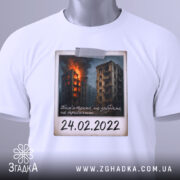 Футболка памʼять 24 лютого, біла, складена на поверхні, фон світлий, принт з палаючими будинками, на футболці є етикетка.