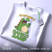 Футболка панікозавр білого кольору, складена на поверхні, з видимим етикеткою, фон світло-сірий, принт з динозавром та написом 'Сьогодні я панікозавр' у яскравих кольорах.