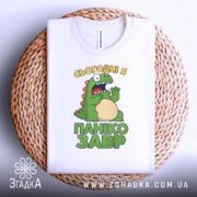 Футболка панікозавр білого кольору, складена на плетеній кошику, фон білий, принт з динозавром та написом 'Сьогодні я панікозавр' у яскравих кольорах.