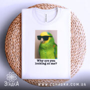 Футболка папуга у окулярах, біла, складена на плетеній кошику, фон світло-сірий, принт з яскравим зеленим папугою в окулярах, текст під ним: 'Why are you looking at me?'.