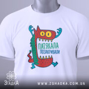 Футболка патякала позакривали, біла, зображена зверху, фон світлий, принт з монстром, який піднімає руки, кольори принта червоний, зелений, жовтий та блакитний, на футболці є етикетка.