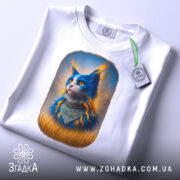 Футболка патріотичний кіт, біла, зображення кота з яскравими синіми і помаранчевими деталями, ракурс зверху, фон світло-сірий, принт з котом у національному вбранні, на футболці є етикетка з зеленим шнурком.