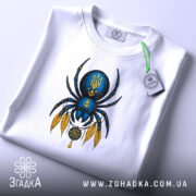 Футболка павук з тризубом, біла, зображена зверху, фон світло-сірий, принт з павуком синього кольору з жовтими деталями, на футболці є етикетка з логотипом.