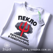Футболка пекло переповнене, біла футболка на плоскій поверхні, з кольоровим принтом, що містить текст "ПЕКЛО ПЕРЕПОВНЕНЕ, Я ПОВЕРНУЛАСЬ" та червоний тризуб на фоні хмар, з етикеткою на зеленій стрічці.