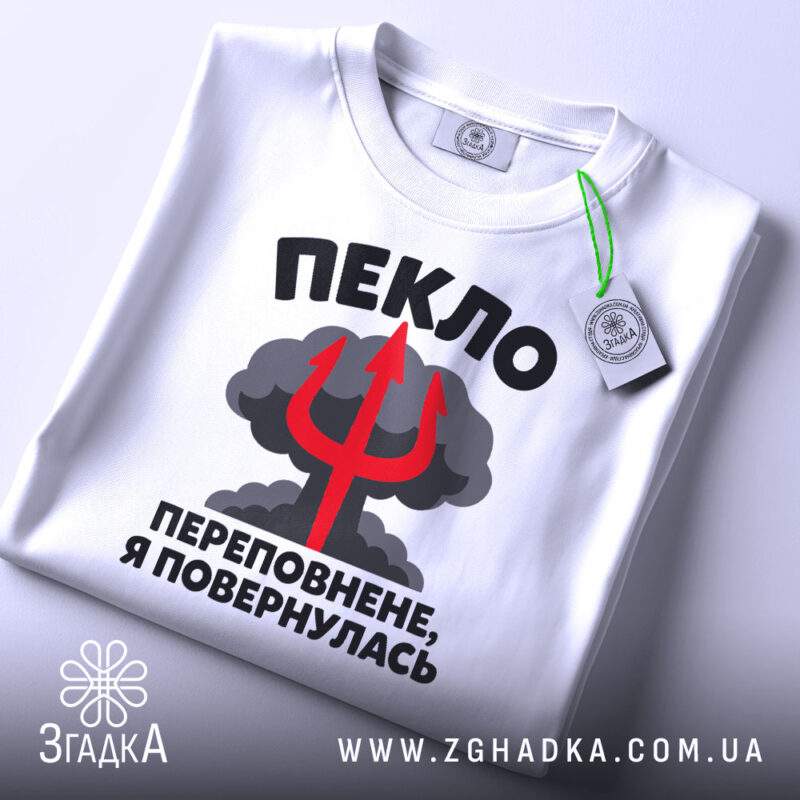 Футболка пекло переповнене, біла футболка на плоскій поверхні, з кольоровим принтом, що містить текст "ПЕКЛО ПЕРЕПОВНЕНЕ, Я ПОВЕРНУЛАСЬ" та червоний тризуб на фоні хмар, з етикеткою на зеленій стрічці.