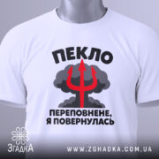 Футболка пекло переповнене, біла футболка на плоскій поверхні, з кольоровим принтом, що містить текст "ПЕКЛО ПЕРЕПОВНЕНЕ, Я ПОВЕРНУЛАСЬ" та червоний тризуб на фоні хмар, з етикеткою на зеленій стрічці.