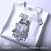 Футболка perfect hero арт білого кольору, складена на поверхні, з видимим етикеткою. Принт зображує персонажа з великими очима та текстом 'PERFECT HERO', на фоні світлого градієнта.