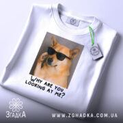 Футболка пес білого кольору, зображена на столі, ракурс зверху, фон світло-сірий, принт з собакою в окулярах та текстом "Why are you looking at me?", на футболці є етикетка.