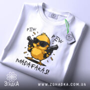 Футболка Pew Pew Madafakas, білого кольору, складена на плоскій поверхні, фон світло-сірий, принт з жовтим курчам у чорних окулярах, яке тримає пістолети, текст 'PEW PEW MADAFAKAS!' під малюнком, на футболці є етикетка з зеленим шнурком.