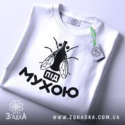 Футболка під мухою білого кольору, зображена зверху, на ній видно чорний принт з малюнком мухи та написом 'під мухою', на комірці є етикетка.