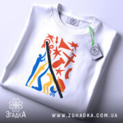 Футболка підняти Україну, біла, зображена на столі, фон світло-сірий, принт кольоровий, з фігурами людей, червоними зірками та іншими елементами, на футболці є етикетка з зеленим шнурком.