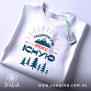 Футболка подорожую отже існую, біла, зображена зверху, на фоні світло-сірого кольору, принт з горами, деревами та текстом, з прикріпленою етикеткою зеленого кольору.