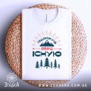 Футболка подорожую отже існую, біла, зображена зверху на плетеному кошику, на фоні світло-сірого кольору, принт містить гори, дерева та текст, з округлим вирізом.