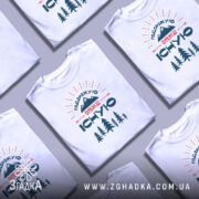 Футболка подорожую отже існую, біла, зображена з правого боку, на фоні світло-сірого кольору, принт містить гори, дерева та текст у стилі ретро.