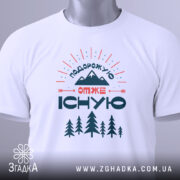 Футболка подорожую отже існую, біла, зображена зверху, на фоні світло-сірого кольору, принт з горами, деревами та текстом, з прикріпленою етикеткою зеленого кольору.