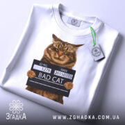 Футболка поганий кіт арешт, біла, зображена зверху, на фоні світлого градієнта, принт з котом у поліцейській табличці з написом 'BAD CAT', з зеленим ярликом.